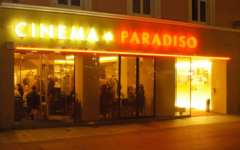 Außenansicht des Cinema Paradiso bei Nacht, mit Menschen, die durch die großen Fenster und die gelbe und rote Leuchtreklame über dem Eingang ins Innere gelangen.