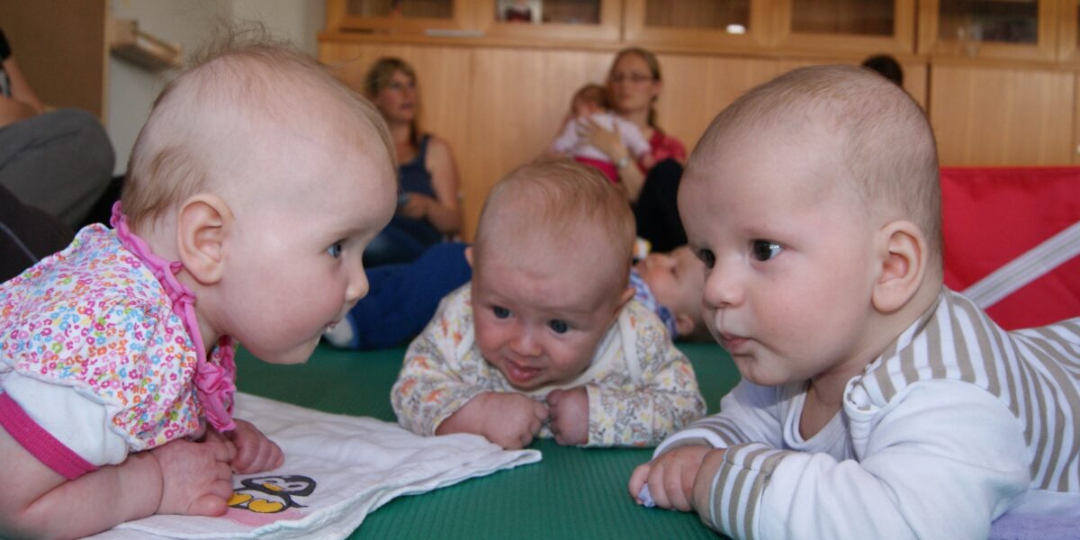 Drei Babys liegen auf dem Bauch, einander zugewandt auf einer grünen Matte, während die Erwachsenen im Hintergrund sitzen.