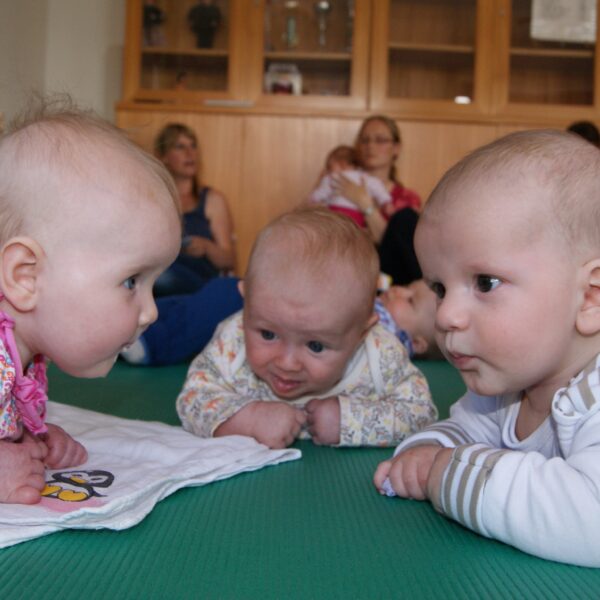 Drei Babys liegen auf dem Bauch, einander zugewandt auf einer grünen Matte, während die Erwachsenen im Hintergrund sitzen.