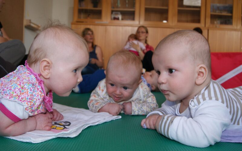 Drei Babys liegen auf dem Bauch, einander zugewandt auf einer grünen Matte, während die Erwachsenen im Hintergrund sitzen.
