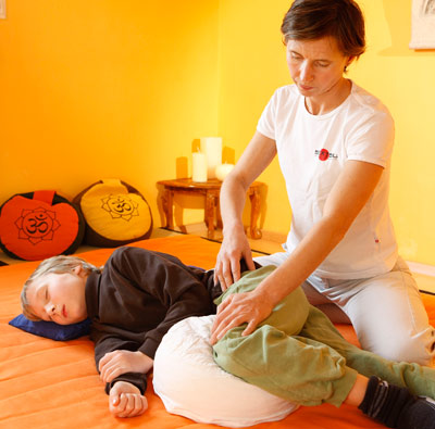 Ein Erwachsener in weißer Kleidung führt eine therapeutische Massage an einem Kind durch, das in einem hell erleuchteten Raum mit orangefarbenem Dekor und dekorativen Kissen auf der Seite liegt.