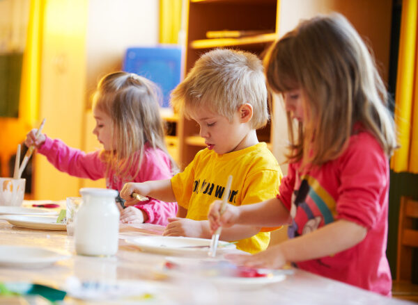 Drei kleine Kinder sitzen an einem Tisch und malen mit Pinseln und Farben in einem hell erleuchteten Klassenzimmer.