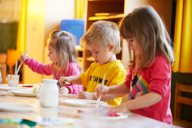 Drei kleine Kinder sitzen an einem Tisch und malen mit Pinseln und Farben in einem hell erleuchteten Klassenzimmer.