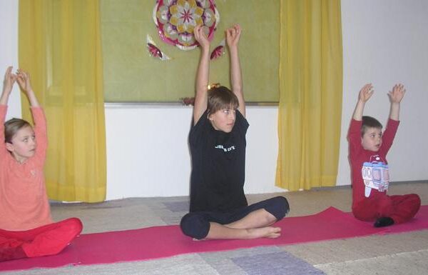 Drei Kinder sitzen im Schneidersitz auf Yogamatten in einem Haus und heben ihre Arme über den Kopf. Im Hintergrund sind gelbe Vorhänge und ein dekorativer Wandbehang zu sehen.