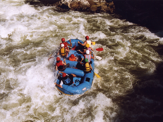 Eine Gruppe von Personen mit Helmen und Schwimmwesten paddelt auf einem blauen Schlauchboot durch Wildwasser-Stromschnellen.