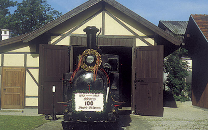 Eine mit einem Kranz geschmückte alte Dampflokomotive steht vor einem offenen Holzschuppen unter einem strahlend blauen Himmel.
