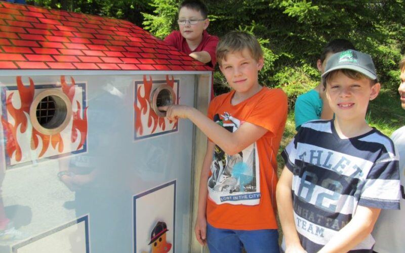 Mehrere Jungen stehen neben einem Modell zur Demonstration des Brandschutzes in Form eines Hauses, wobei ein Junge seine Hand in eine kreisförmige Öffnung an der Seite steckt.