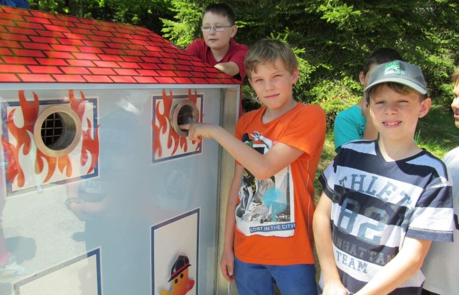 Mehrere Jungen stehen neben einem Modell zur Demonstration des Brandschutzes in Form eines Hauses, wobei ein Junge seine Hand in eine kreisförmige Öffnung an der Seite steckt.