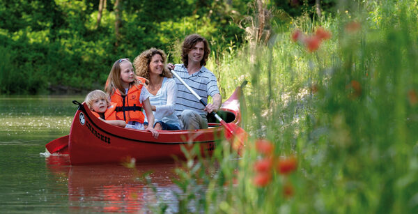 Eine vierköpfige Familie paddelt in einem roten Kanu auf einem ruhigen Fluss, umgeben von Grün und Blumen, mit zwei Kindern in orangefarbenen Schwimmwesten.