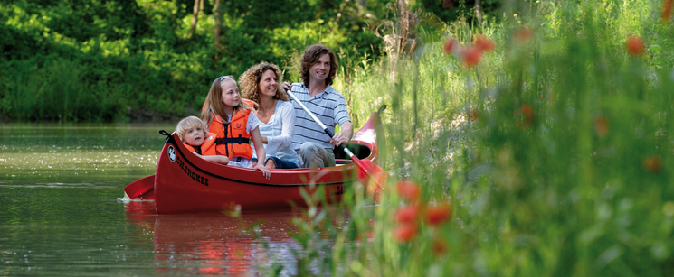 Eine vierköpfige Familie paddelt in einem roten Kanu auf einem ruhigen Fluss, umgeben von Grün und Blumen, mit zwei Kindern in orangefarbenen Schwimmwesten.
