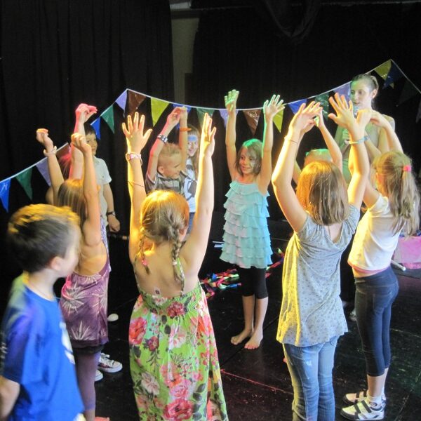 Eine Gruppe von Kindern steht mit erhobenen Armen im Kreis und nimmt an einer Aktivität auf einer mit bunten Wimpeln geschmückten Bühne teil.