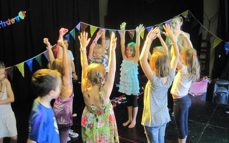Eine Gruppe von Kindern steht mit erhobenen Armen im Kreis und nimmt an einer Aktivität auf einer mit bunten Wimpeln geschmückten Bühne teil.