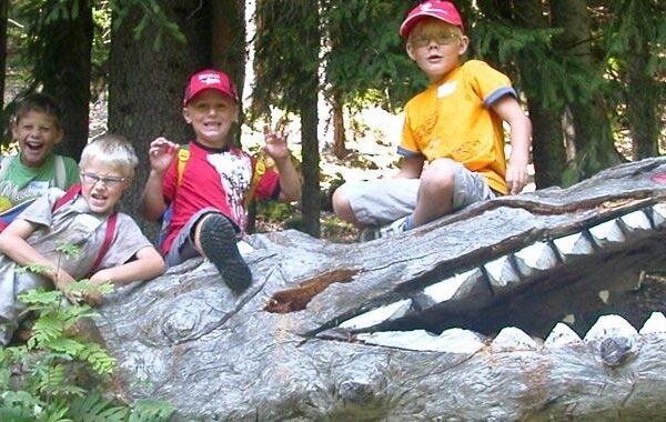 Eine Gruppe von Kindern sitzt in Pose auf einer großen hölzernen Krokodilskulptur in einem Waldgebiet, lächelt und trägt Rucksäcke und Hüte.