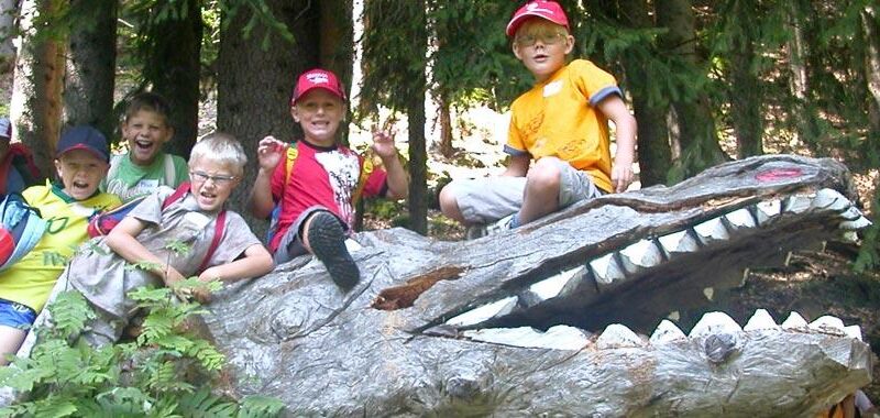 Eine Gruppe von Kindern sitzt in Pose auf einer großen hölzernen Krokodilskulptur in einem Waldgebiet, lächelt und trägt Rucksäcke und Hüte.