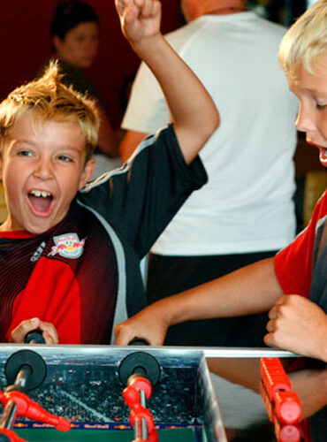Zwei Jungen spielen Tischfußball; der eine jubelt mit erhobener Faust und aufgeregtem Gesichtsausdruck, während der andere auf den Tisch blickt.
