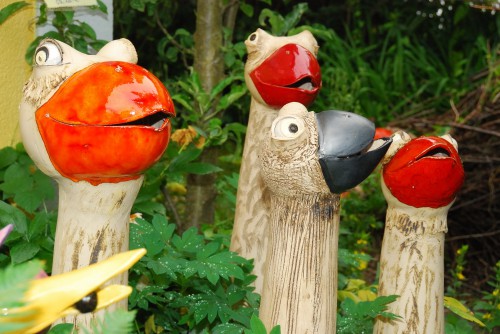Vier keramische Vogelskulpturen mit farbenfrohen, glänzenden Schnäbeln stehen inmitten von Grünpflanzen in einem Garten im Freien.