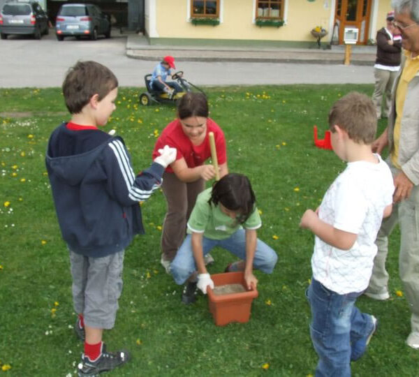 Eine Gruppe von Kindern und ein Erwachsener sitzen draußen im Gras. Ein Kind hält einen Stock über einen mit Erde gefüllten Topf, während die anderen zusehen. Im Hintergrund sind ein Kinderwagen und ein Gebäude zu sehen.