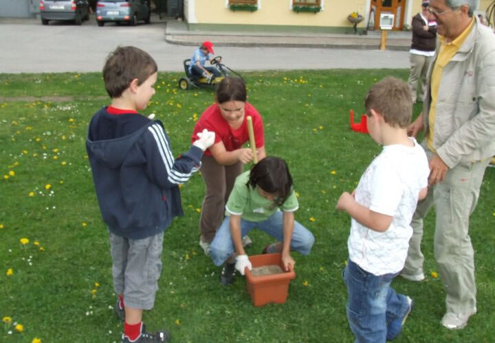 Eine Gruppe von Kindern und ein Erwachsener sitzen draußen im Gras. Ein Kind hält einen Stock über einen mit Erde gefüllten Topf, während die anderen zusehen. Im Hintergrund sind ein Kinderwagen und ein Gebäude zu sehen.