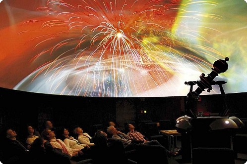 In einem Planetarium sitzende Personen sehen eine farbenfrohe, feurige Weltraumprojektion auf einer großen Kuppel, wobei im Vordergrund eine Projektionsmaschine zu sehen ist.