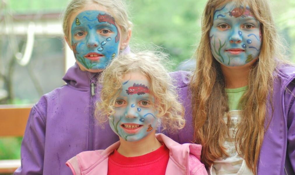 Drei Kinder mit blauer Gesichtsbemalung und Unterwassermotiven stehen zusammen im Freien, alle tragen lila oder rosa Jacken.
