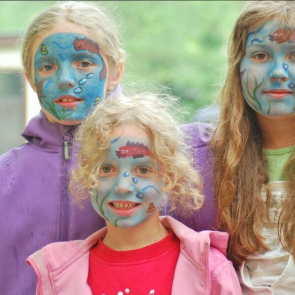 Drei Kinder mit blauer Gesichtsbemalung und Unterwassermotiven stehen zusammen im Freien, alle tragen lila oder rosa Jacken.
