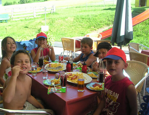 Sechs Kinder sitzen um einen Tisch im Freien, essen Pommes frites und trinken Limonade, während im Hintergrund eine Spielplatzrutsche und eine Wiese zu sehen sind.