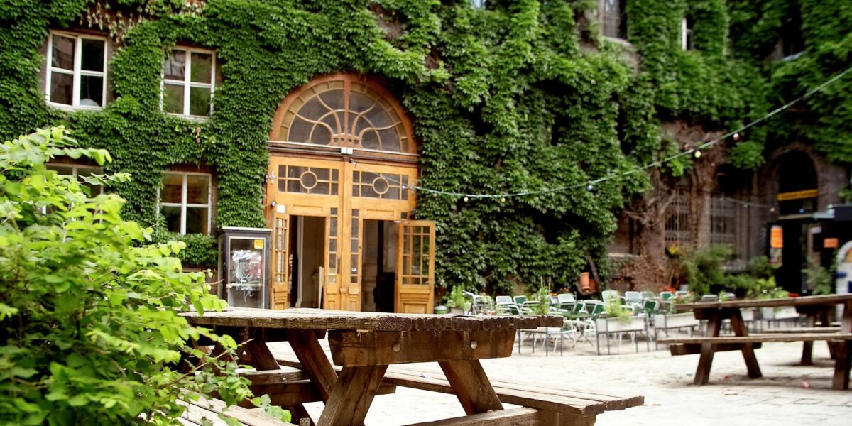 Hölzerne Picknicktische in einem Innenhof mit einem großen efeubewachsenen Gebäude und einem gewölbten Holztor im Hintergrund; leere Stühle und Lichterketten sind zu sehen.
