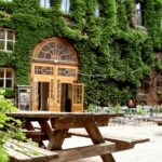 Hölzerne Picknicktische in einem Innenhof mit einem großen efeubewachsenen Gebäude und einem gewölbten Holztor im Hintergrund; leere Stühle und Lichterketten sind zu sehen.