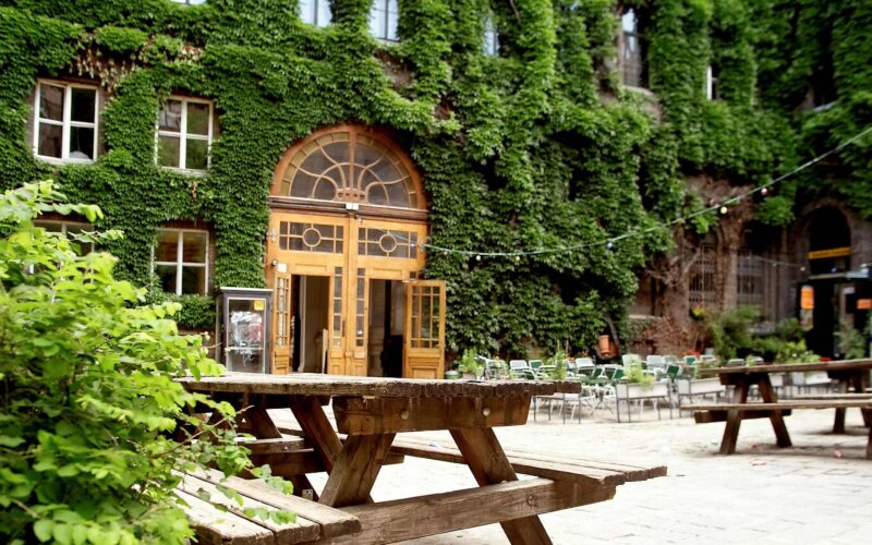 Hölzerne Picknicktische in einem Innenhof mit einem großen efeubewachsenen Gebäude und einem gewölbten Holztor im Hintergrund; leere Stühle und Lichterketten sind zu sehen.