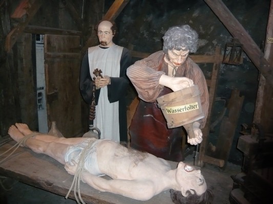 In einem historischen Wachsfigurenkabinett simulieren zwei Figuren die Wasserfolter an einem gefesselten Menschen, der auf einem Holztisch liegt; ein Eimer mit der Aufschrift "Wasserfolter" wird auf sein Gesicht geschüttet.