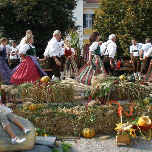 Menschen in traditioneller Kleidung tanzen im Kreis auf einer mit Stroh bedeckten Plattform; ein Mädchen sitzt auf einem großen Kürbis im Vordergrund, umgeben von Herbstdekorationen.