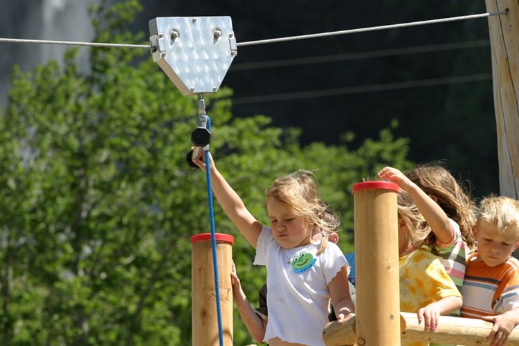 Mehrere Kinder warten auf einer hölzernen Plattform, während ein Mädchen sich darauf vorbereitet, an einem sonnigen Tag im Freien eine Seilrutsche zu benutzen.