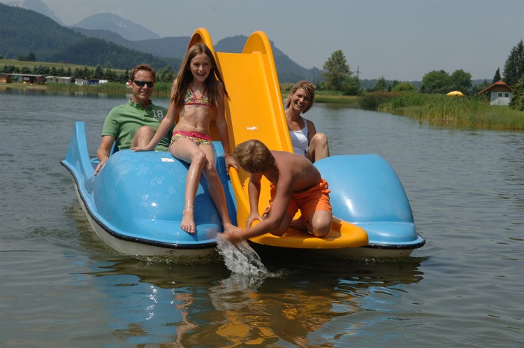 Eine vierköpfige Familie sitzt in einem blauen Paddelboot mit gelber Rutsche auf einem See; der Junge vorne greift ins Wasser, während die anderen lächeln.
