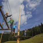 Vier Personen mit Helmen und Klettergurten navigieren in einem Hochseilgarten im Freien mit blauem Himmel, grasbewachsenen Hügeln und Bäumen im Hintergrund.
