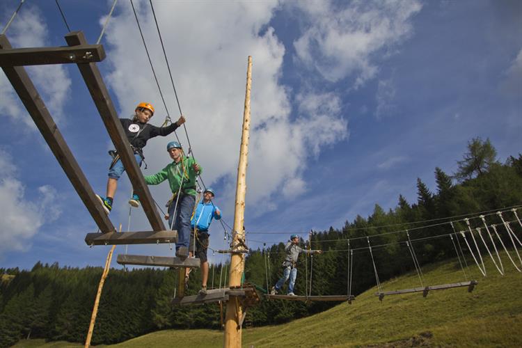 Vier Personen mit Helmen und Klettergurten navigieren in einem Hochseilgarten im Freien mit blauem Himmel, grasbewachsenen Hügeln und Bäumen im Hintergrund.