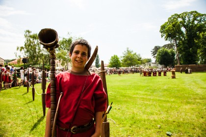 Ein Junge in einem historischen Kostüm hält ein Requisiten-Horn und steht auf einer Wiese während einer Nachstellung im Freien mit Zuschauern im Hintergrund.