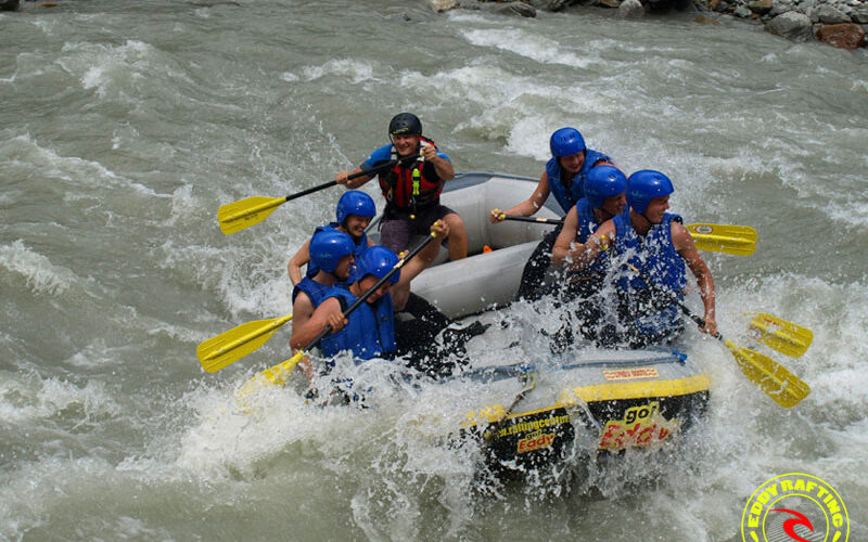 Sechs Personen mit blauen Helmen und Schwimmwesten beim Wildwasser-Rafting auf einem rauen Fluss, die durch das spritzende Wasser paddeln. In der unteren rechten Ecke ist ein Logo für "Sport Rafting" zu sehen.