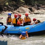 Eine Gruppe von Personen mit Helmen und Schwimmwesten fährt in einem blauen Schlauchboot einen Fluss hinunter, wobei mehrere Teilnehmer ins Wasser fallen.