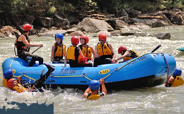 Eine Gruppe von Personen mit Helmen und Schwimmwesten fährt in einem blauen Schlauchboot einen Fluss hinunter, wobei mehrere Teilnehmer ins Wasser fallen.