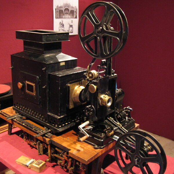 Ein alter Filmprojektor mit großen Rollen und Messingbeschlägen steht auf einem Tisch vor einer roten Wand, mit einem Schwarz-Weiß-Foto im Hintergrund.