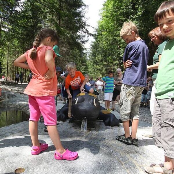 Kinder stehen herum und spielen in der Nähe einer schwarzen Froschstatue mit Wasserspeiern in einem Park, der von Bäumen und anderen Besuchern umgeben ist.