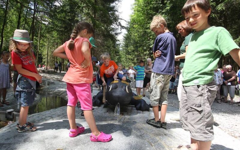 Kinder stehen herum und spielen in der Nähe einer schwarzen Froschstatue mit Wasserspeiern in einem Park, der von Bäumen und anderen Besuchern umgeben ist.