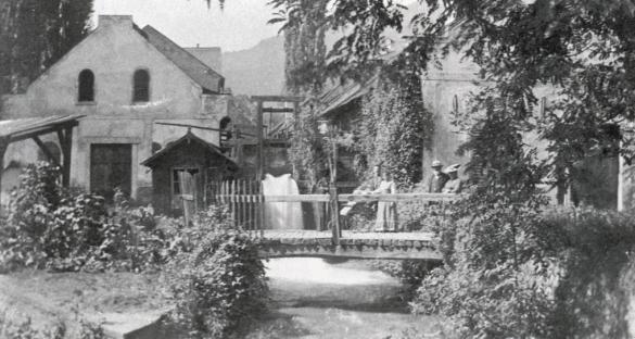 Schwarz-Weiß-Foto einer kleinen Brücke über einen Bach, auf der mehrere Personen sitzen, sowie alte Gebäude, die von Bäumen und Laub im Hintergrund umgeben sind.