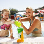 Ein Erwachsener und ein Kind spielen an einem sonnigen Tag am Rande eines Freibads mit Wasserspielzeug.