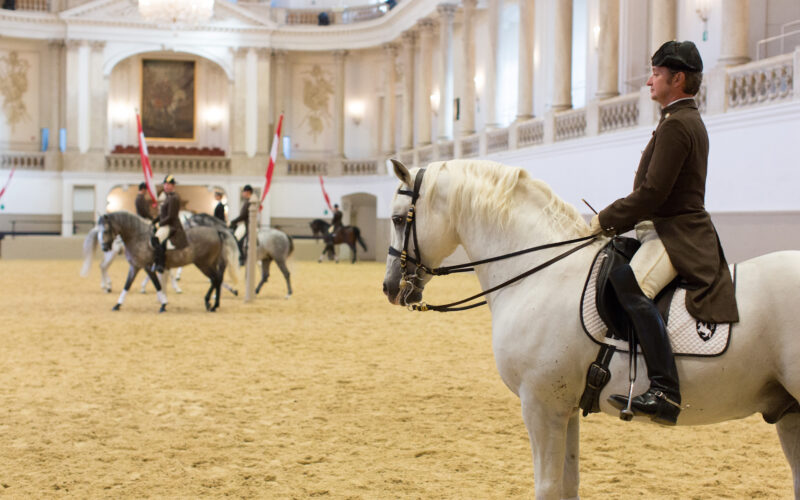 Reiter in Uniform üben Dressur auf Pferden in einer prächtigen Reithalle mit hohen Decken und Kronleuchtern.