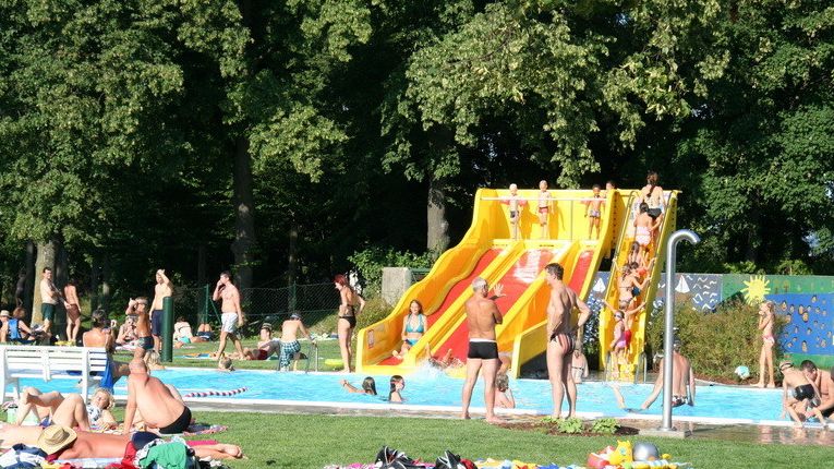 Menschen an einem Freibad mit einer gelben und roten Wasserrutsche, umgeben von Bäumen und Gras, die einen sonnigen Tag genießen.
