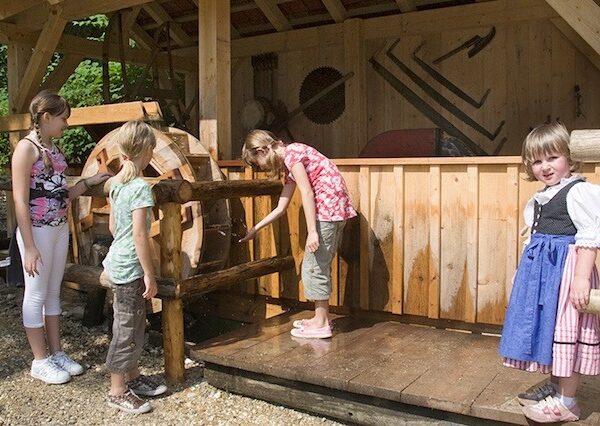 Vier junge Mädchen stehen im Freien um ein hölzernes Wasserrad, wobei ein Mädchen ein traditionelles Kleid trägt.