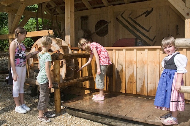 Vier junge Mädchen stehen im Freien um ein hölzernes Wasserrad, wobei ein Mädchen ein traditionelles Kleid trägt.