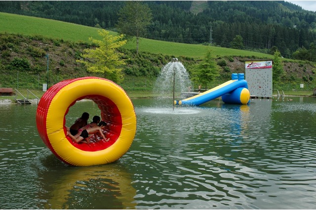 Kinder sitzen in einem großen aufblasbaren gelben und roten Rad, das auf einem Teich schwimmt, während im Hintergrund ein Springbrunnen, eine Wasserrutsche und eine Kletterwand zu sehen sind.