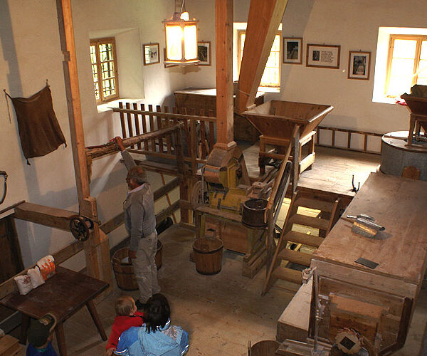 Das Innere einer traditionellen Wassermühle mit hölzernen Maschinen, Eimern, Balken und Besuchern, die die Geräte beobachten. Durch die Fenster fällt natürliches Licht ein.